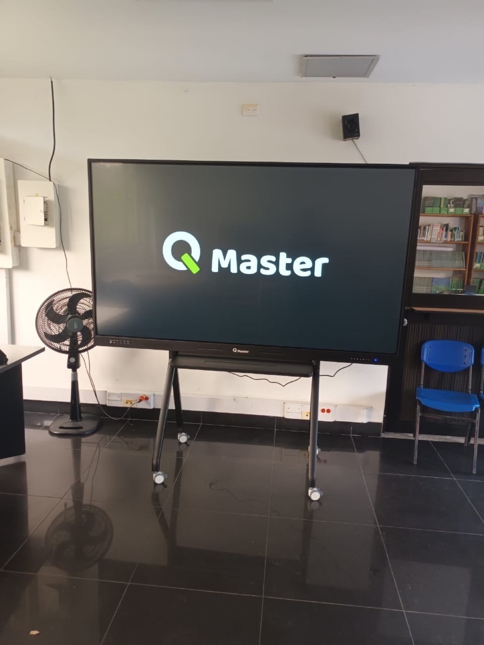 Pantalla interactiva QMaster en soporte móvil junto a una estantería