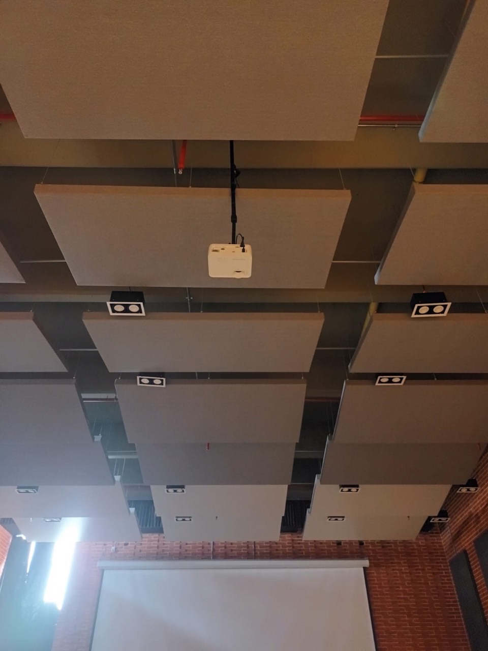 Videoproyector suspendido entre paneles acústicos de un auditorio escolar, con el telón al fondo